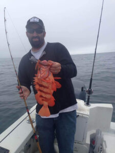 Lucky angler holds up a beautiful Tiger Rockfish he just caught with Prisoner Rock Charters on the Black Cadillac. A great California fishing trip! The man has a goatee, dark hat, and dark sunglasses.