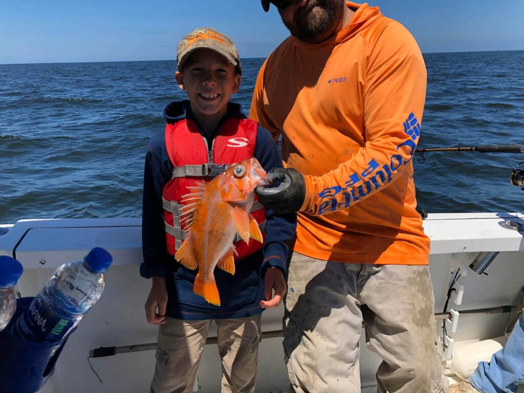Captain Jason holds a canary rockfish up next to the young angler who just caught it. The young angler is smiling and wearing a hat and lifejacket.